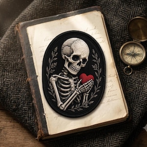 May include: An oval embroidered patch featuring a white human skull and ribcage holding a small, bright red heart. The skeleton is framed by delicate, thin white leafy branches on a black background, creating a detailed, high-contrast design.