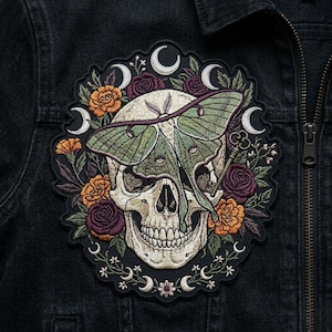 May include: Embroidered patch featuring a skull with a green moth, surrounded by orange and burgundy flowers, crescent moons, and a key. The patch is on a black denim jacket.