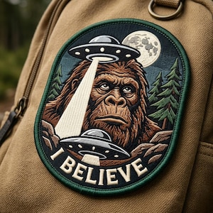 May include: Embroidered patch featuring Bigfoot being beamed up by a UFO. The patch has a green border and the words "I BELIEVE" in white. The background includes a full moon and pine trees. The patch is attached to a tan backpack.