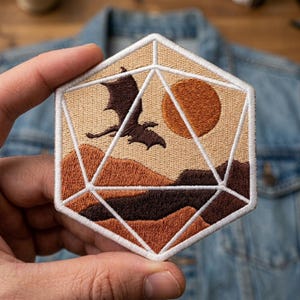 May include: Embroidered patch in the shape of a D20 die, featuring a dragon silhouette flying over a mountain landscape with a sun. The patch has a beige background with brown and orange details, and white geometric lines.