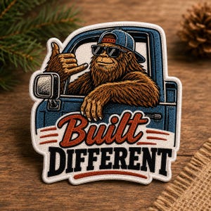 May include: An embroidered patch featuring a brown Bigfoot character wearing a blue baseball cap and black sunglasses, leaning out of a blue truck window while making a shaka hand gesture. The text reads Built DIFFERENT in red and black.