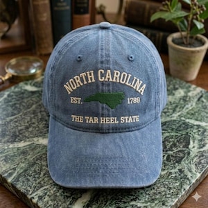 Puede incluir: Una gorra de b&eacute;isbol de mezclilla azul con texto bordado que dice North Carolina, Est. 1789 y The Tar Heel State, junto con un contorno verde del estado de Carolina del Norte centrado en el panel frontal de la gorra.