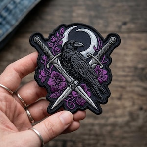 May include: Embroidered patch featuring a black raven perched on crossed swords, with a crescent moon and purple floral accents. The patch is black with white and purple details, suitable for jackets, bags, or other fabric items.