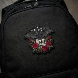 May include: Black backpack with a decorative patch featuring a skull, roses, and a bat with stars. The patch has a gothic aesthetic with red, white, and black colors. The backpack is made of a textured, durable material.