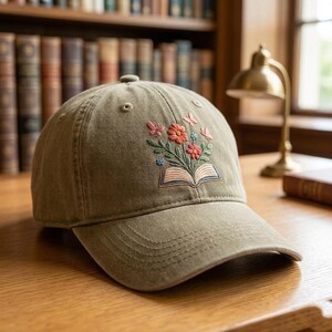 May include: An olive green baseball cap featuring an embroidered design of an open book with a bouquet of red, pink, and blue flowers emerging from the pages, accented by small butterflies, with a curved brim and stitched eyelets on the crown.