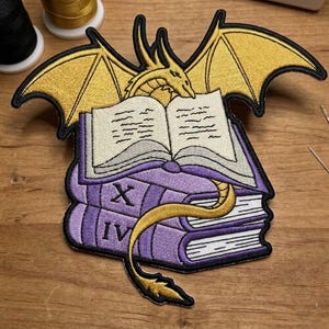 May include: Embroidered patch featuring a gold dragon atop an open book, with purple books stacked below. The dragon has outstretched wings and a long tail that wraps around the books. The patch has black borders.