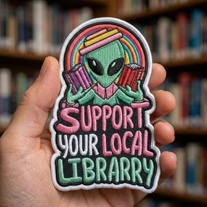 May include: Embroidered patch featuring a green alien wearing a pink hat and holding books, set against a rainbow. The patch reads "SUPPORT YOUR LOCAL LIBRARRY" in pink and green lettering, with a white border.