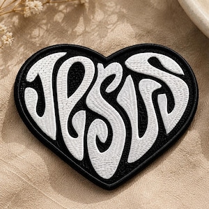 May include: A heart-shaped embroidered patch featuring a black background with the word JESUS stitched in bold, white, stylized lettering. The patch has a thick black border and a textured surface created by the dense white and black thread work.
