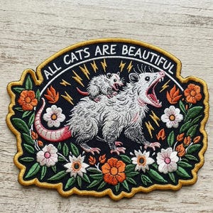 May include: Embroidered patch featuring a white opossum with an open mouth, carrying a smaller opossum on its back. The patch is surrounded by flowers and leaves, with the text "ALL CATS ARE BEAUTIFUL" in a curved banner.
