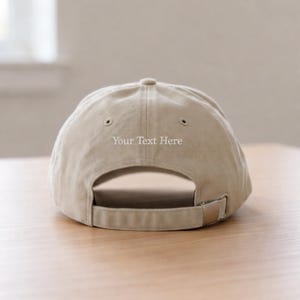 Puede incluir: Una gorra de b&eacute;isbol de color beige con una correa ajustable y una hebilla de metal en la parte posterior. La tela es de un color tostado claro s&oacute;lido y el texto "Your Text Here" est&aacute; bordado en hilo blanco sobre la abertura trasera.