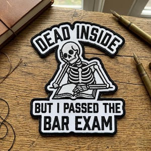 May include: A black and white patch featuring a skeleton reading a book, with the text "DEAD INSIDE" above and "BUT I PASSED THE BAR EXAM" below. The patch has a white border and is on a wooden surface.
