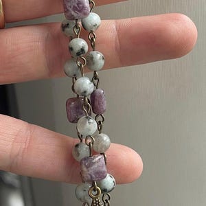 May include: A rosary with round and square beads in shades of gray, white, and purple. The beads are strung on a chain with a metal cross and a medallion featuring a religious figure.