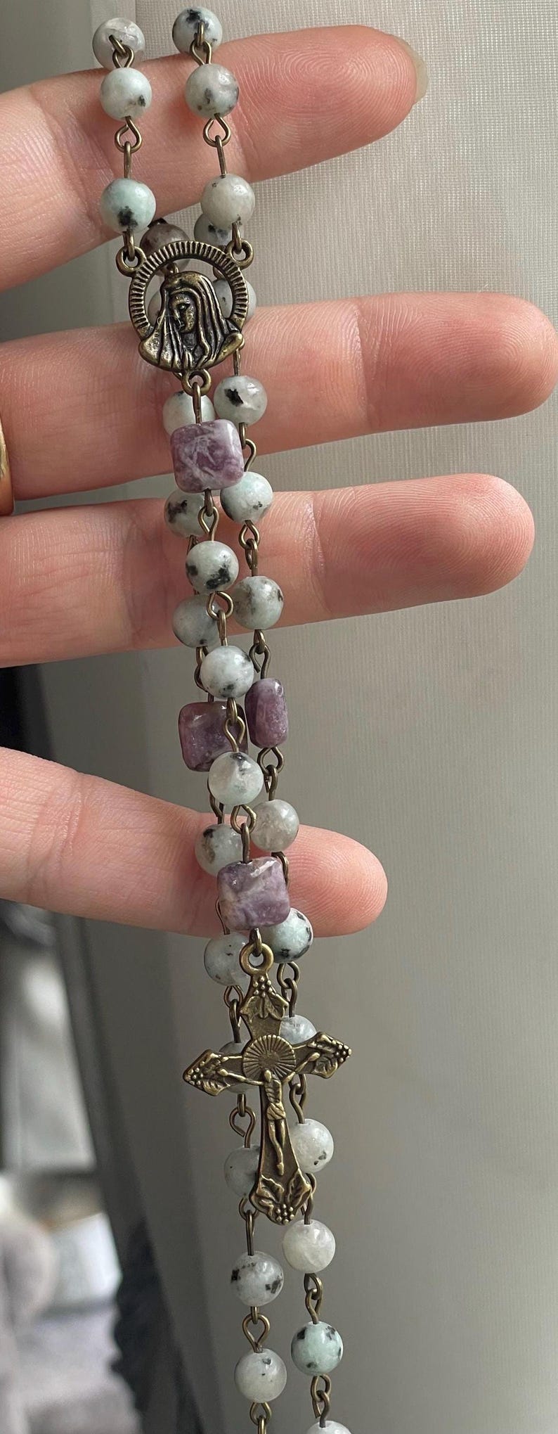 May include: A rosary with round and square beads in shades of gray, white, and purple. The beads are strung on a chain with a metal cross and a medallion featuring a religious figure.