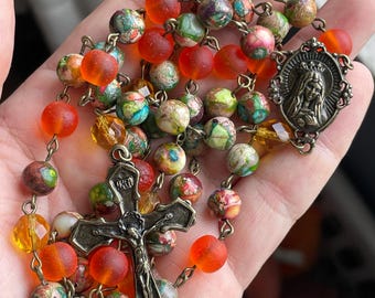 May include: A rosary with colorful beads in shades of orange, red, green, and white. The rosary features a metal crucifix and a pendant with a religious figure. The beads are strung together with metal links.