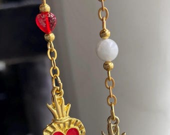 May include: Gold-coloured earrings with a chain design. One earring features a red heart pendant, while the other has a black heart pendant. Both have a gold crown design. The earrings also have various beads in red, white, and pink.