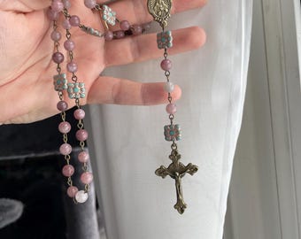May include: A rosary with pink gemstone beads and decorative floral accents. The rosary features a crucifix and a central medallion with a religious image. The metal components have a bronze finish.