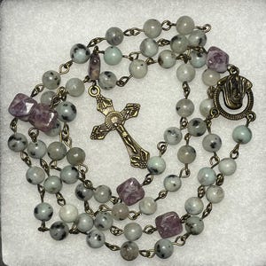 May include: A rosary with light green and gray speckled beads, interspersed with purple square beads. A detailed bronze cross and clasp are featured. The beads are strung on bronze-colored chain.