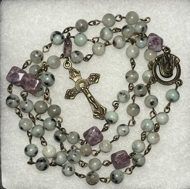 May include: A rosary with light green and gray speckled beads, interspersed with purple square beads. A detailed bronze cross and clasp are featured. The beads are strung on bronze-colored chain.