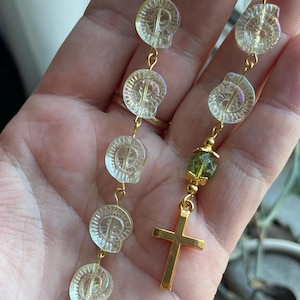 May include: A rosary with clear, iridescent skull-shaped beads and gold-colored accents. The rosary features a gold cross pendant and a gold oval pendant with a religious figure. The beads are connected by gold wire.