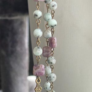 May include: A rosary with a bronze cross and a pendant featuring a religious figure. The beads are a mix of round and square shapes in shades of green, white, and purple. The rosary is made with a gold-colored chain.