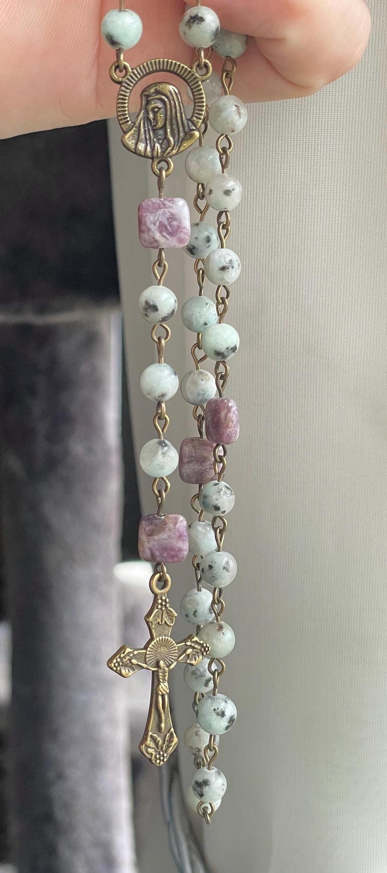 May include: A rosary with a bronze cross and a pendant featuring a religious figure. The beads are a mix of round and square shapes in shades of green, white, and purple. The rosary is made with a gold-colored chain.