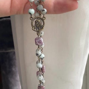 May include: A rosary with light green and white speckled beads, interspersed with purple square beads. The rosary features a bronze-colored crucifix and a medallion with a religious figure.