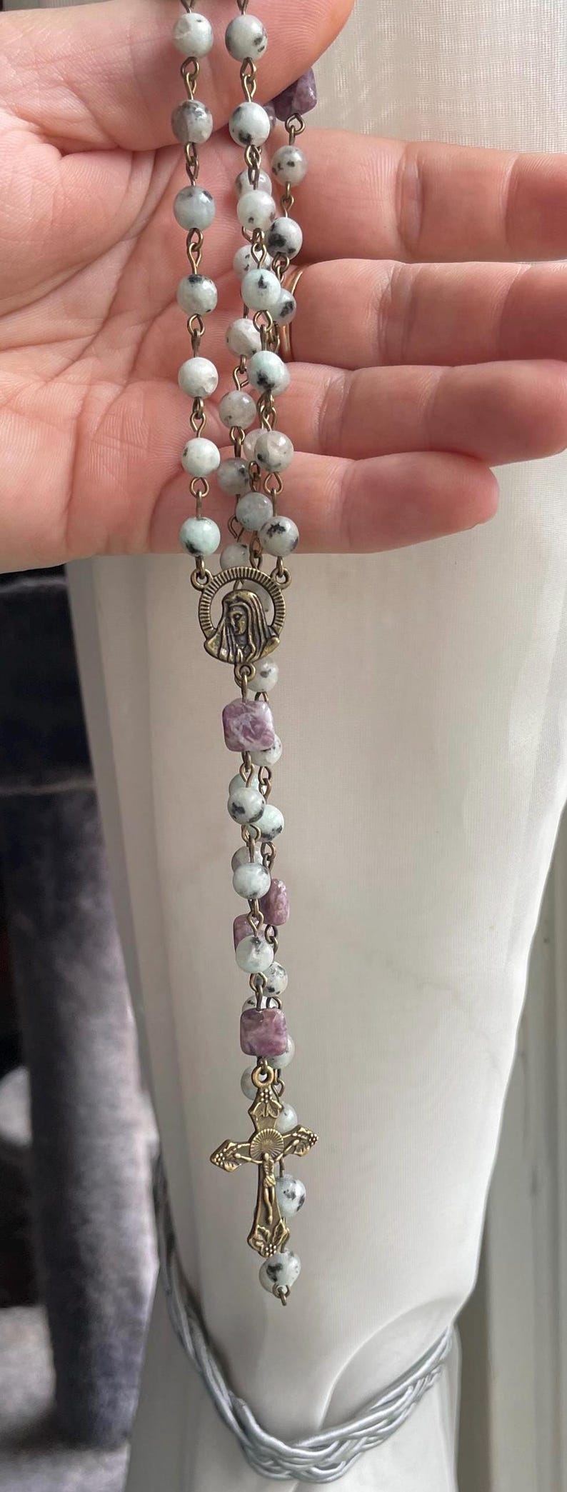 May include: A rosary with light green and white speckled beads, interspersed with purple square beads. The rosary features a bronze-colored crucifix and a medallion with a religious figure.
