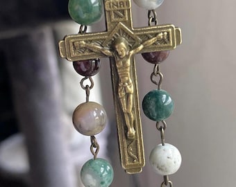 May include: A rosary with green, white, and maroon stone beads. The beads are connected by gold-colored metal links. A gold-colored metal crucifix with a figure of Jesus Christ hangs from the rosary.