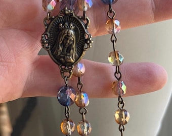 May include: A rosary with iridescent, faceted beads in shades of amber, blue, and purple. The metal chain and centerpiece have an antique bronze finish. The centerpiece features a religious image.