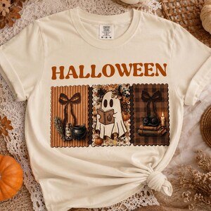 Puede incluir: Una camiseta de manga corta color crema con la palabra HALLOWEEN en letras naranjas sobre tres paneles rectangulares. Los paneles muestran un lazo con una botella y un caldero, un fantasma leyendo un libro y un gato negro con una vela.