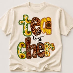 Puede incluir: Una camiseta de manga corta color crema con un estampado gráfico que incluye el texto "tea Best cher". Las letras están decoradas con patrones de girasoles amarillos y estampados de leopardo marrones, con la palabra "Best" en letra negra.