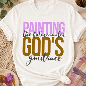 Puede incluir: Una camiseta blanca de manga corta con el texto "PAINTING the future under GOD'S guidance" impreso en la parte delantera. La palabra "PAINTING" es de color morado claro, "the future under" y "guidance" son gris oscuro y "GOD'S" es marrón.
