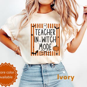 Puede incluir: Una camiseta de manga corta color marfil con un estampado gráfico de rayas verticales naranjas, texto negro que dice "TEACHER IN WITCH MODE" e ilustraciones de dos pequeños fantasmas blancos, una calabaza, murciélagos negros y estrellas.