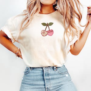 Puede incluir: Una camiseta de manga corta color crema con un gráfico central de dos cerezas. Las cerezas presentan una estética de patchwork, mostrando patrones florales, de lunares y de cuadros en tonos rosa, rojo y verde.