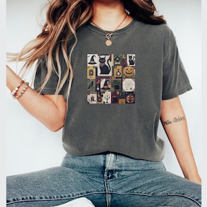 Puede incluir: Una camiseta de manga corta en color gris carbón con un estampado gráfico cuadrado en el centro. El diseño consiste en una cuadrícula de pequeñas ilustraciones que incluyen gatos negros, calabazas, sombreros de bruja, velas y frascos.