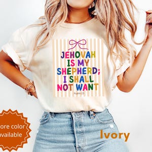 Puede incluir: Una camiseta de manga corta color marfil con un estampado gráfico de un lazo rosa sobre rayas verticales beige y el texto multicolor: JEHOVAH IS MY SHEPHERD; I SHALL NOT WANT, con el texto Psalm 23 impreso debajo de la frase principal.