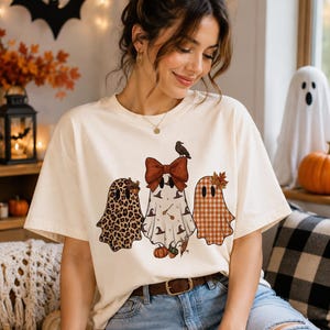 Puede incluir: Una camiseta de manga corta color crema con un gráfico de tres fantasmas. El fantasma de la izquierda tiene estampado de leopardo, el del centro lleva un lazo rojo grande y patrones de sombreros de bruja, y el de la derecha tiene un patrón de cuadros naranja y blanco.