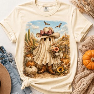 Puede incluir: Una camiseta de manga corta color crema con un estampado gráfico de un fantasma que lleva un sombrero de vaquero con lazo, botas de vaquero marrones y sostiene un ramo de flores. La escena incluye calabazas, cactus y pájaros.