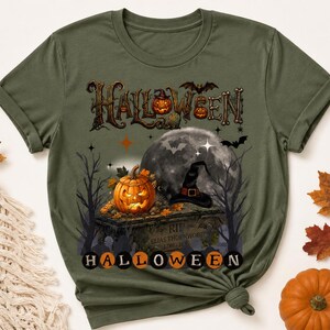 Puede incluir: Una camiseta verde oliva con un estampado gráfico de una calabaza brillante sobre una lápida, un sombrero de bruja, una luna llena, árboles desnudos y murciélagos. El texto HALLOWEEN aparece en la parte superior e inferior en círculos naranjas.