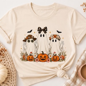Puede incluir: Una camiseta de color crema con un gráfico de tres fantasmas. El fantasma de la izquierda lleva sombrero y gafas de sol, el del centro un lazo negro y el de la derecha una corona de flores. Están rodeados de calabazas naranjas y flores.