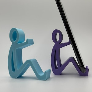 May include: Two stylized phone stands shaped like seated figures. One is light blue, the other is purple, and holds a black smartphone. The stands are designed to prop up a phone for hands-free viewing.