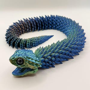 Peut inclure: Un jouet serpent articulé bleu et vert. Le jouet est composé de nombreux petits segments texturés qui lui permettent de bouger et de se plier. La tête du serpent est détaillée avec un œil réaliste et une bouche ouverte.