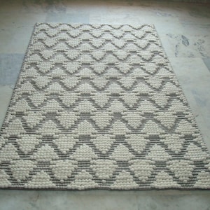 May include: Rectangular rug with a repeating geometric pattern. The rug features a cream-coloured design with grey accents and a grey border. The pattern consists of zig-zag shapes. The rug is made of a textured material.