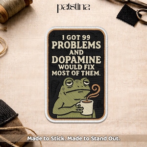 May include: A rectangular embroidered patch with a black background and a tan border. It features a green frog holding a white coffee mug with steam rising, and the text: I GOT 99 PROBLEMS AND DOPAMINE WOULD FIX MOST OF THEM.