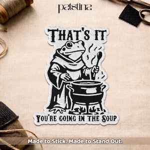 May include: Embroidered patch featuring a frog in a monk's robe stirring a cauldron. The patch reads "THAT'S IT" above the frog and "YOU'RE GOING IN THE SOUP" below. The patch is white with black details and the text "Made to Stick. Made to Stand Out."