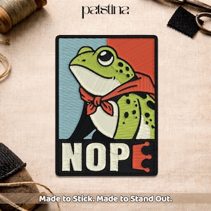 May include: Embroidered patch featuring a green frog wearing a red bandana, set against a blue and red background. The word "NOPE" is embroidered in white, with an orange "E". The patch is rectangular with a black border.