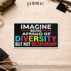 May include: Black rectangular patch with a white border. The patch reads "IMAGINE BEING AFRAID OF DIVERSITY BUT NOT DICTATORSHIP" in white, red, and rainbow-coloured text. The patch is on a beige fabric.