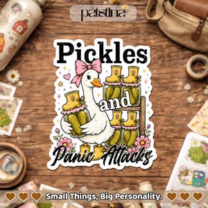 May include: A whimsical sticker featuring a white goose wearing a pink bow and yellow boots, holding a jar of pickles. The text reads "Pickles and Panic Attacks" with floral accents. The sticker is on a wooden surface.