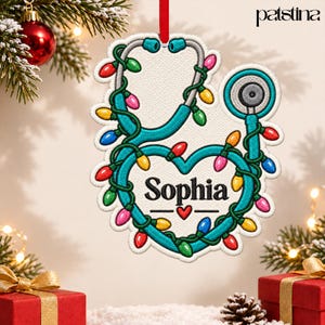 May include: A white, heart-shaped ornament featuring a teal stethoscope wrapped in colorful Christmas lights. The name Sophia is printed in black text in the center, accompanied by a small red heart. The ornament hangs from a red ribbon.