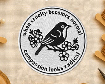 May include: Embroidered patch with a black bird perched on a flowering branch, surrounded by the text "When cruelty becomes normal, compassion looks radical." The patch is circular with a white background and black border.